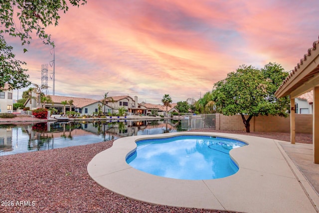 pool at dusk with a patio area, fence, an outdoor pool, and a water view