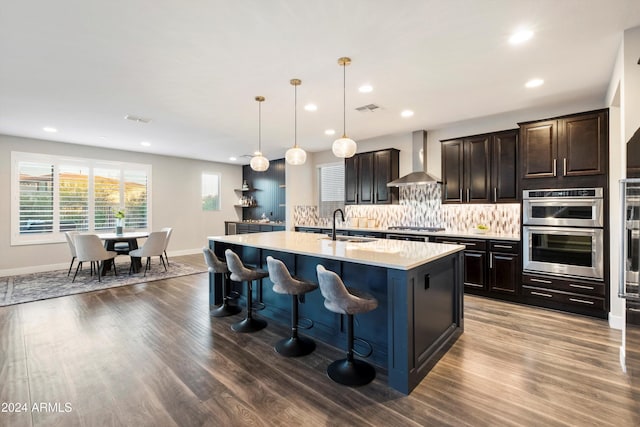 kitchen with sink, stainless steel appliances, wall chimney range hood, an island with sink, and pendant lighting