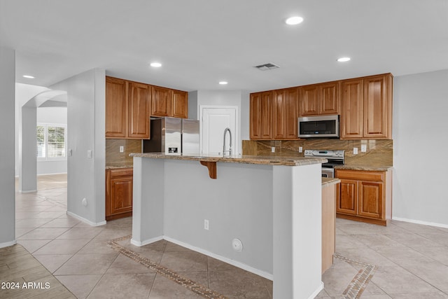 kitchen with decorative backsplash, appliances with stainless steel finishes, a center island with sink, and light stone counters