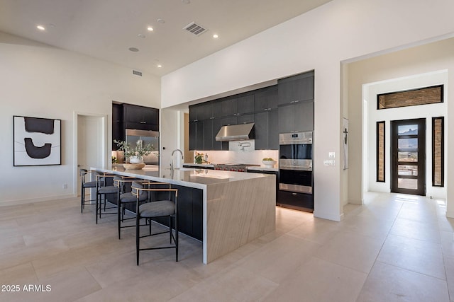 kitchen with a kitchen island with sink, a towering ceiling, a kitchen breakfast bar, stainless steel appliances, and decorative backsplash