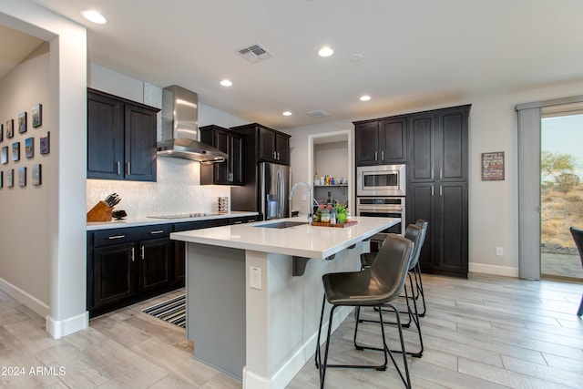 kitchen featuring wall chimney exhaust hood, an island with sink, a breakfast bar, appliances with stainless steel finishes, and light wood-type flooring