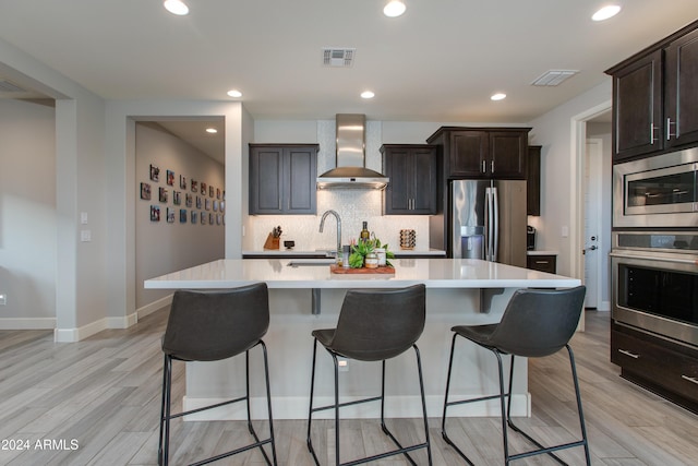 kitchen featuring appliances with stainless steel finishes, wall chimney exhaust hood, a kitchen island with sink, sink, and light hardwood / wood-style flooring