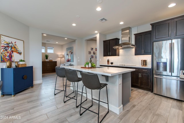 kitchen with a center island with sink, wall chimney range hood, light hardwood / wood-style flooring, stainless steel fridge, and a kitchen bar