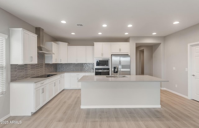 kitchen with wall chimney range hood, an island with sink, light wood-type flooring, stainless steel appliances, and visible vents