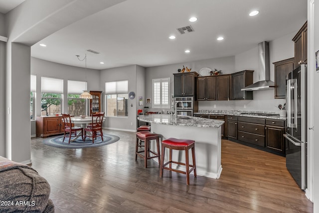 kitchen featuring plenty of natural light, appliances with stainless steel finishes, dark wood-type flooring, wall chimney range hood, and an island with sink