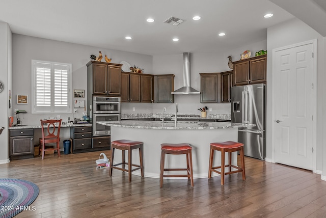 kitchen featuring light stone counters, appliances with stainless steel finishes, wall chimney exhaust hood, wood-type flooring, and a kitchen island with sink