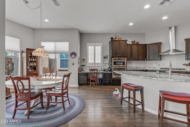 kitchen featuring wall chimney exhaust hood, double oven, dark hardwood / wood-style flooring, light stone counters, and dark brown cabinetry