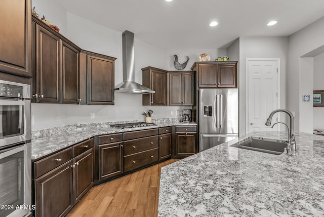 kitchen with dark brown cabinetry, appliances with stainless steel finishes, sink, wall chimney range hood, and light wood-type flooring