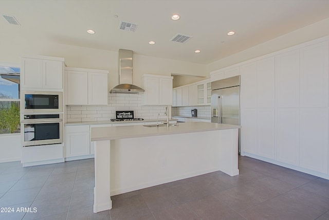 kitchen featuring white cabinets, an island with sink, built in appliances, wall chimney exhaust hood, and backsplash