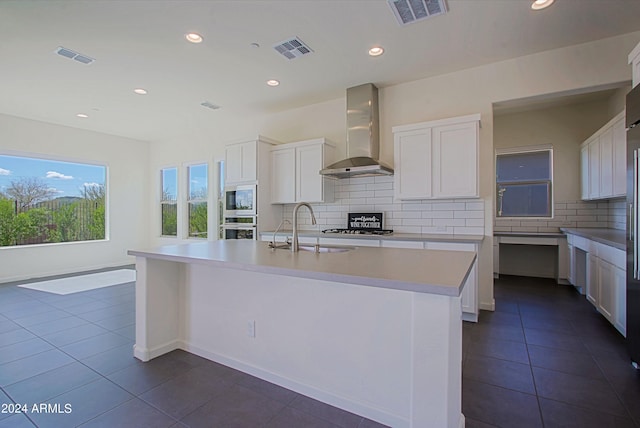 kitchen featuring appliances with stainless steel finishes, dark tile floors, white cabinetry, a center island with sink, and wall chimney exhaust hood