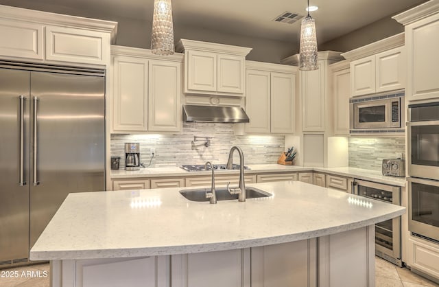 kitchen with a kitchen island with sink, hanging light fixtures, built in appliances, wine cooler, and light stone countertops