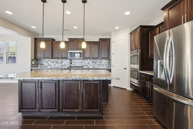 kitchen with light stone countertops, stainless steel appliances, an island with sink, and pendant lighting