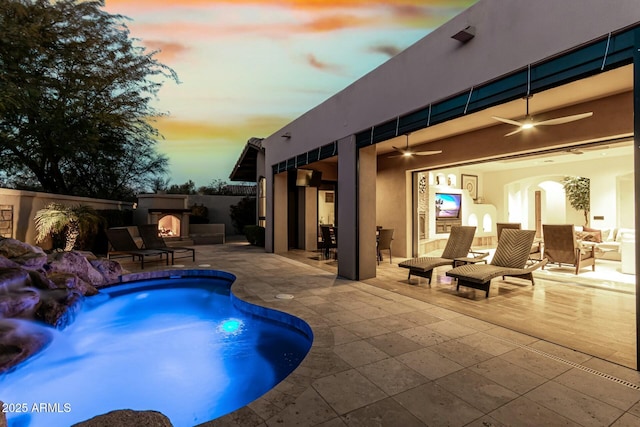 pool at dusk with a warm lit fireplace, a patio area, fence, and a ceiling fan