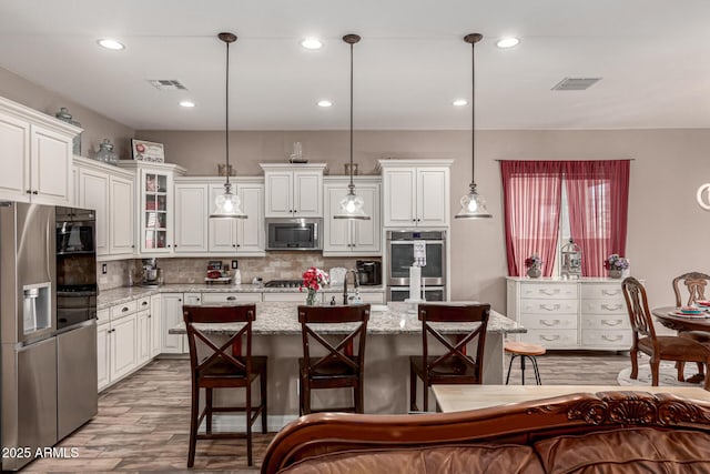 kitchen with a kitchen island with sink, stainless steel appliances, light stone countertops, glass insert cabinets, and pendant lighting