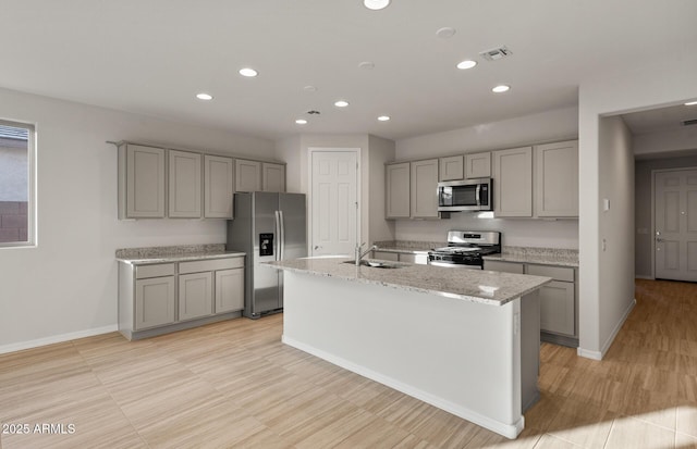 kitchen with sink, a center island with sink, gray cabinets, stainless steel appliances, and light stone countertops