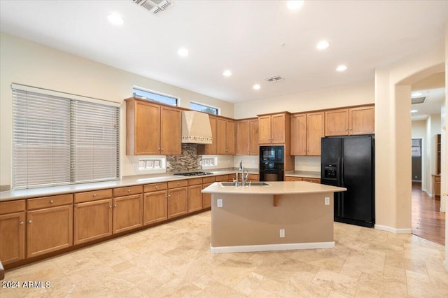 kitchen with custom range hood, tasteful backsplash, black appliances, a kitchen island with sink, and sink