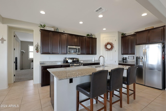 kitchen featuring sink, light stone counters, an island with sink, dark brown cabinets, and appliances with stainless steel finishes