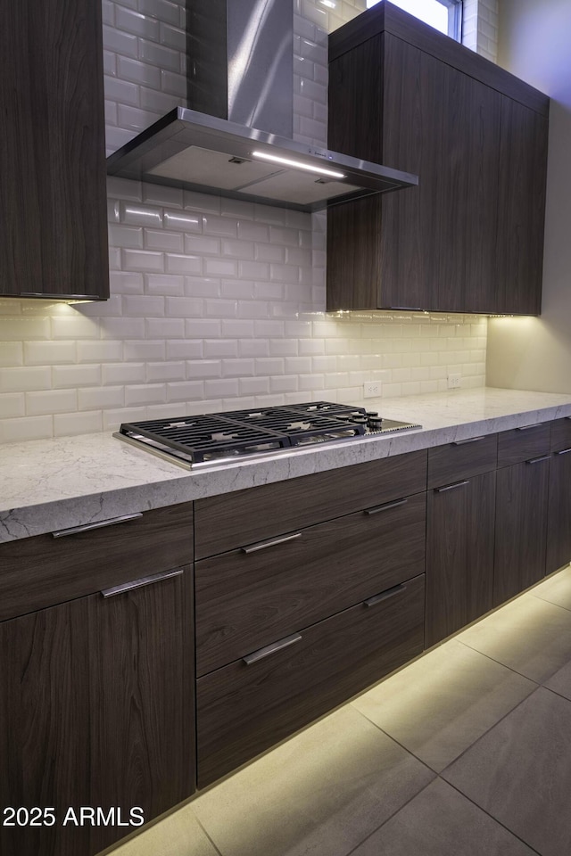 kitchen featuring backsplash, dark brown cabinetry, stainless steel gas cooktop, light tile patterned flooring, and wall chimney exhaust hood