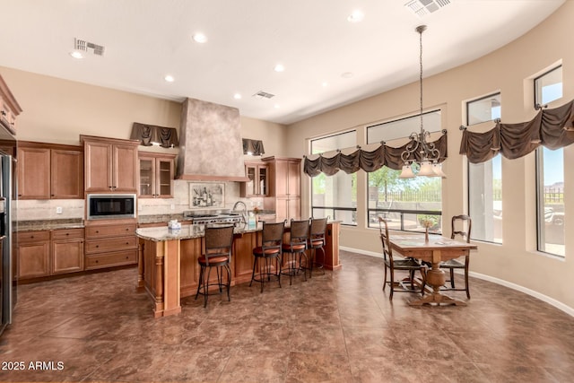 kitchen with built in microwave, a kitchen island with sink, custom exhaust hood, brown cabinets, and glass insert cabinets
