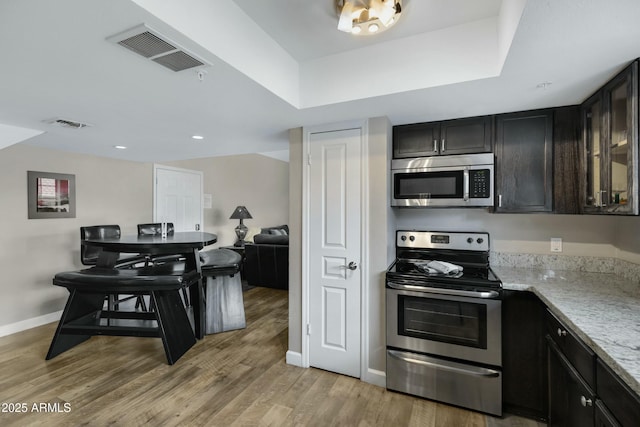 kitchen with appliances with stainless steel finishes, dark brown cabinets, light stone counters, a tray ceiling, and light hardwood / wood-style floors