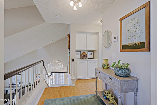 hall featuring a textured ceiling, an upstairs landing, light wood-style flooring, and lofted ceiling