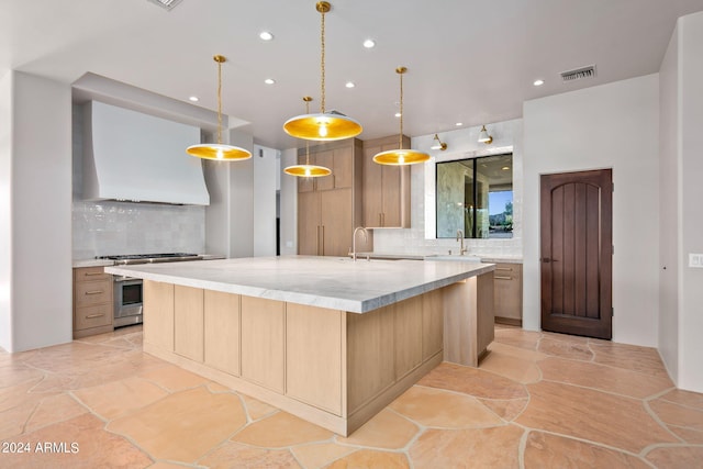 kitchen with decorative light fixtures, stainless steel range with gas stovetop, custom exhaust hood, a kitchen island with sink, and backsplash