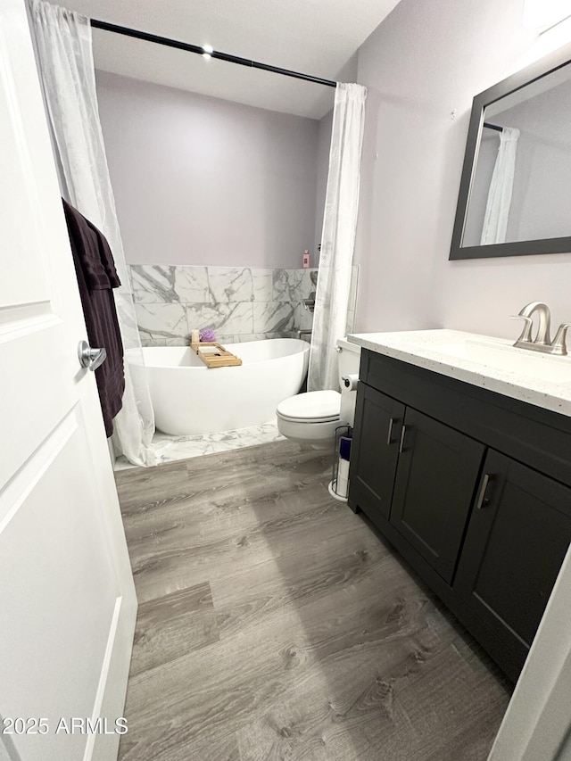 full bathroom featuring toilet, a freestanding bath, wood finished floors, and vanity