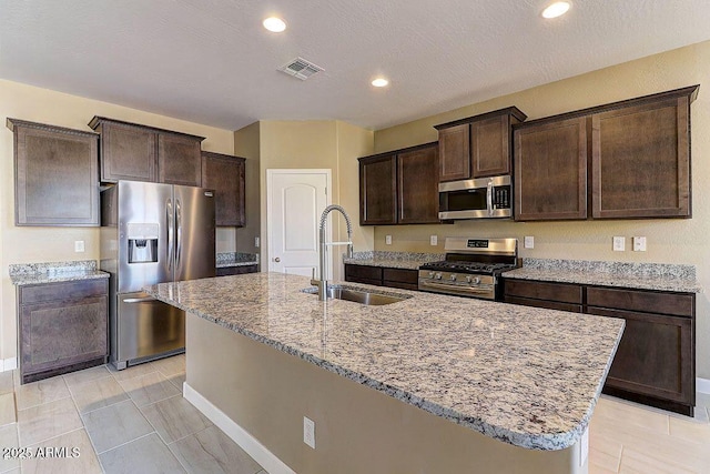 kitchen featuring visible vents, appliances with stainless steel finishes, a kitchen island with sink, a sink, and dark brown cabinets