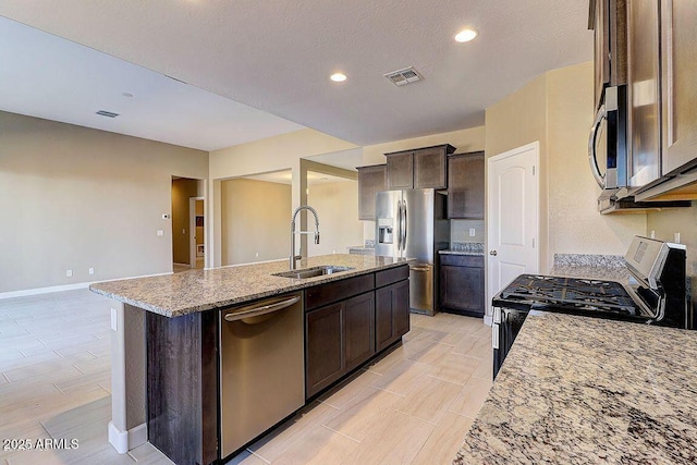 kitchen with appliances with stainless steel finishes, a kitchen island with sink, a sink, dark brown cabinetry, and light stone countertops