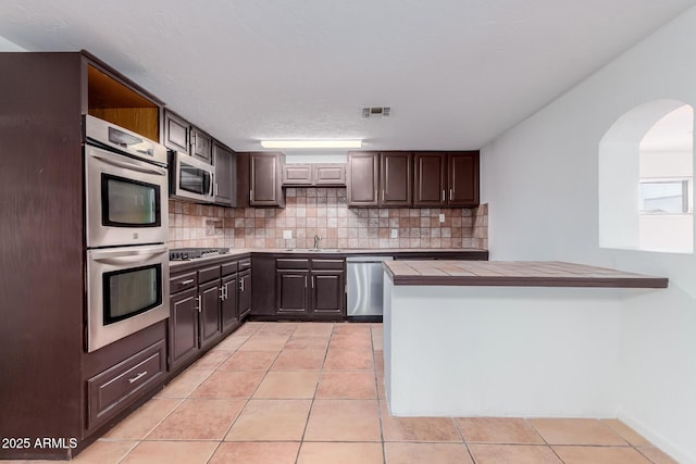 kitchen with tile counters, dark brown cabinetry, tasteful backsplash, light tile patterned floors, and appliances with stainless steel finishes