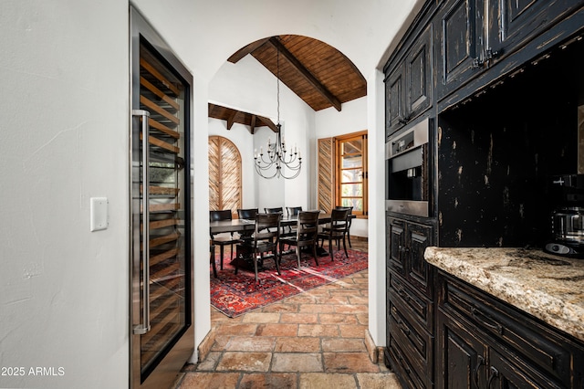kitchen with wooden ceiling, vaulted ceiling with beams, light stone counters, dark cabinetry, and wine cooler