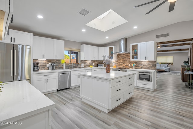 kitchen with white cabinets, vaulted ceiling with skylight, a kitchen island, wall chimney range hood, and stainless steel appliances