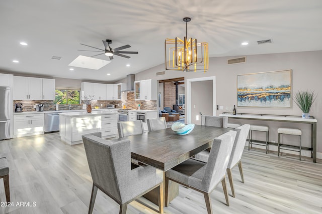 dining area featuring ceiling fan with notable chandelier, a wood stove, light wood-type flooring, vaulted ceiling with skylight, and sink