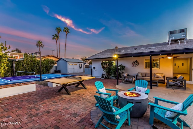 patio terrace at dusk featuring a storage shed, tennis court, and an outdoor living space with a fire pit