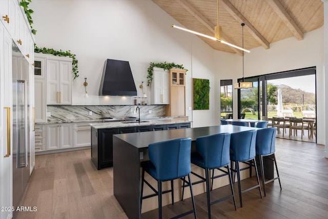 kitchen featuring high vaulted ceiling, backsplash, a center island with sink, and range hood