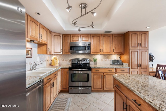 kitchen with appliances with stainless steel finishes, light stone counters, a tray ceiling, sink, and light tile patterned floors