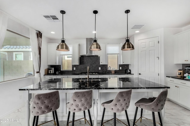 kitchen featuring white cabinetry, a kitchen island with sink, wall chimney range hood, and decorative light fixtures