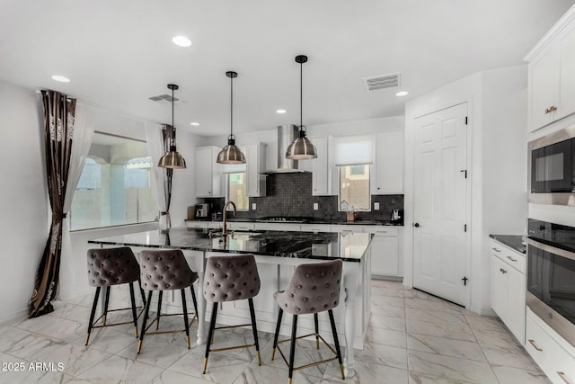 kitchen featuring wall chimney exhaust hood, pendant lighting, oven, a kitchen island with sink, and white cabinets