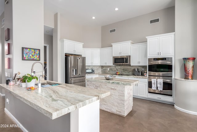 kitchen with a towering ceiling, appliances with stainless steel finishes, sink, white cabinetry, and a center island with sink