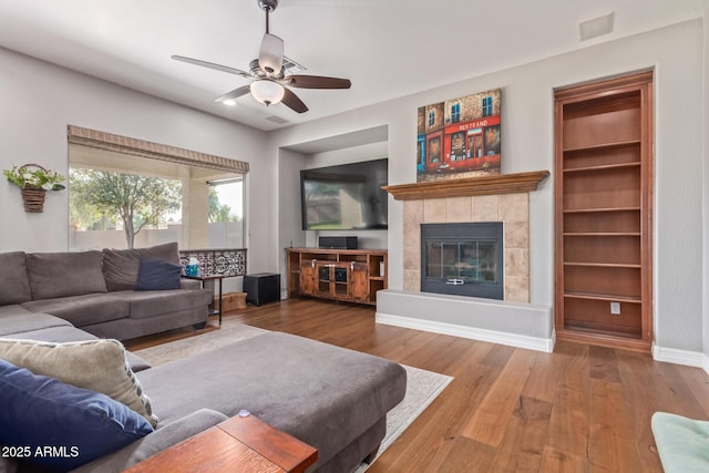 living room with a tiled fireplace, hardwood / wood-style floors, ceiling fan, and built in shelves