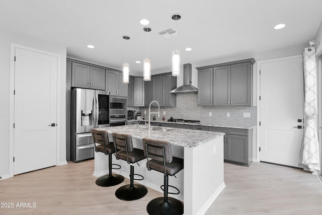 kitchen featuring visible vents, gray cabinetry, stainless steel appliances, wall chimney range hood, and light stone countertops