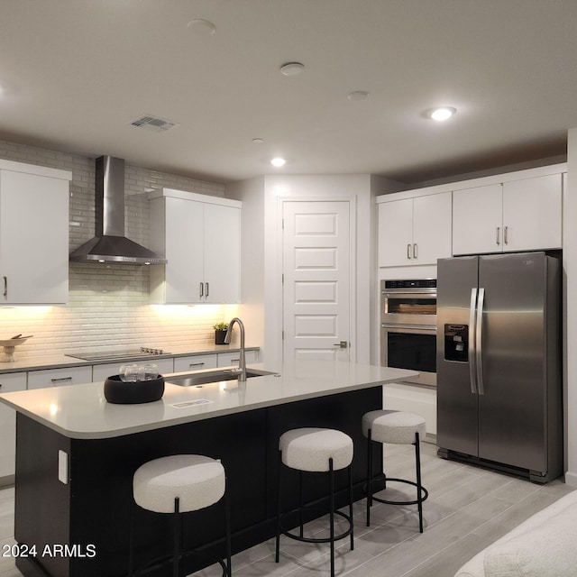 kitchen with wall chimney exhaust hood, stainless steel appliances, sink, a center island with sink, and white cabinetry