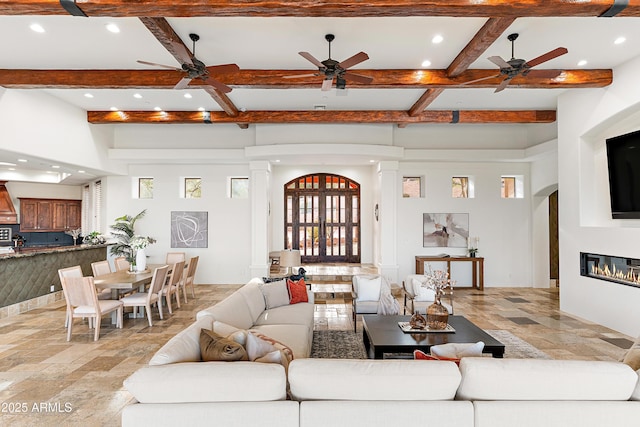 living area featuring ornate columns, stone tile floors, beamed ceiling, and a glass covered fireplace