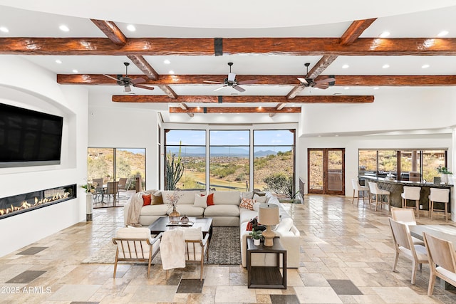 living room featuring ceiling fan, recessed lighting, beam ceiling, a glass covered fireplace, and stone tile flooring