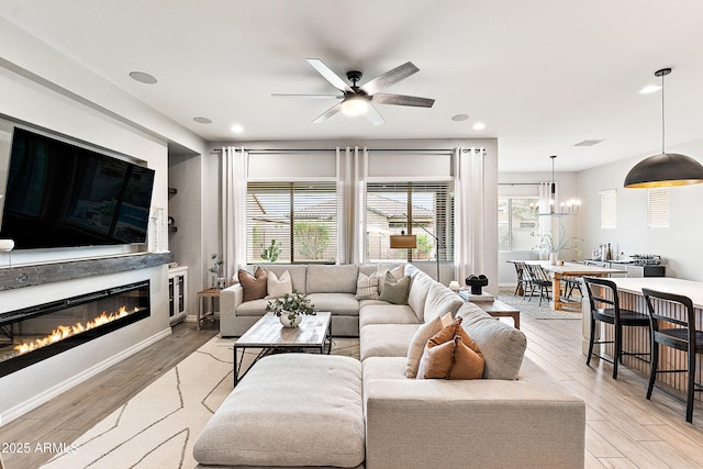 living room with a glass covered fireplace, ceiling fan with notable chandelier, light wood-type flooring, and a wealth of natural light