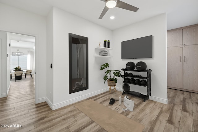 exercise area featuring light wood-style floors, a fireplace, baseboards, and ceiling fan with notable chandelier