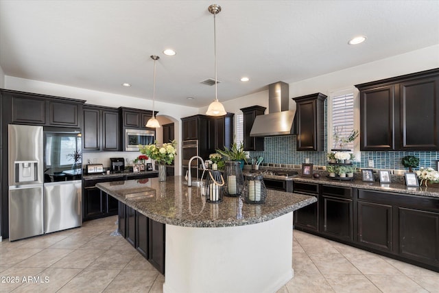 kitchen with appliances with stainless steel finishes, a wealth of natural light, pendant lighting, a kitchen island with sink, and wall chimney range hood