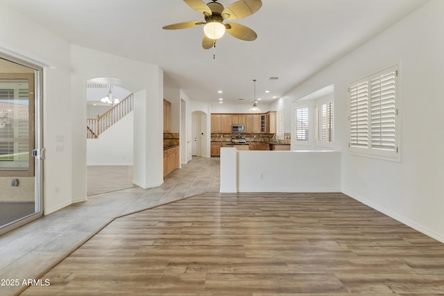 unfurnished living room featuring arched walkways, ceiling fan, baseboards, stairway, and light wood-type flooring