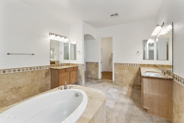 full bathroom featuring a garden tub, tile walls, two vanities, visible vents, and a sink