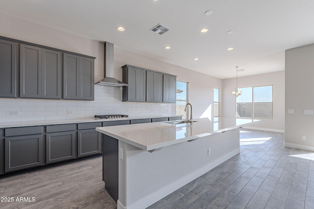 kitchen with backsplash, wall chimney exhaust hood, sink, an inviting chandelier, and an island with sink